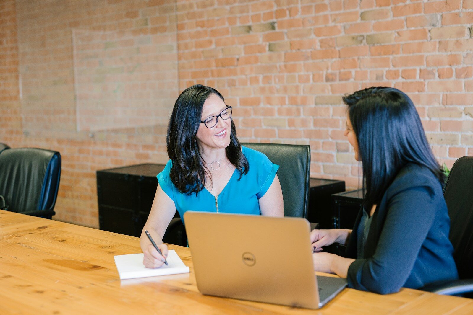 Two women engage in a friendly discussion at a conference table, with a laptop and notepad in front of them, highlighting collaborative work.