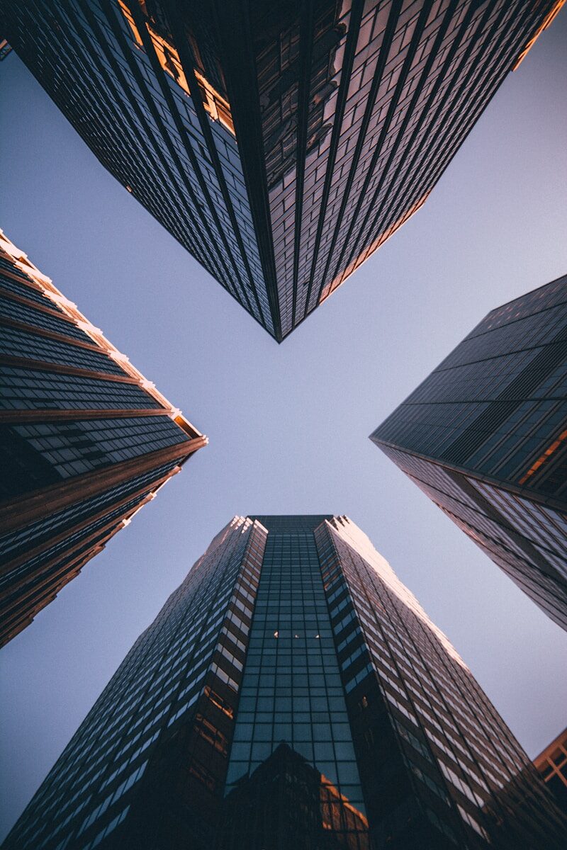 View upward from ground level, capturing the dramatic angles of towering glass skyscrapers against a clear blue sky.